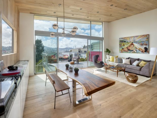 Open-plan contemporary interior combining a kitchen, dining area and living room, with extensive floor-to-ceiling glazing opening toward a balcony and mountainous urban landscape. The room features a warm timber ceiling and flooring, timber kitchen cabinetry, a live-edge dining table, mid-century-inspired seating, a grey sofa, and a sculptural multi-light pendant.