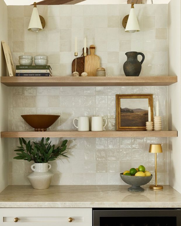 A warm, carefully styled kitchen shelving vignette with three open timber shelves against a glossy cream square-tile backsplash. The shelves display ceramics, wooden boards, books, a framed landscape artwork, a vase with greenery, a fruit bowl and a small brass lamp. A pale stone benchtop, white cabinetry and a black appliance are visible below.