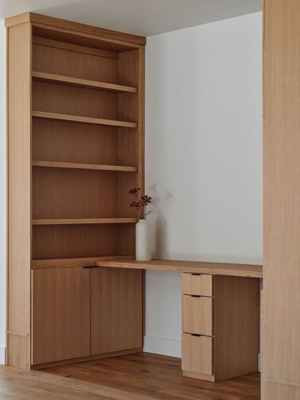 A warm, minimalist built-in timber study nook featuring full-height open shelving, lower storage cabinetry, and an integrated desk with drawers. The joinery is set against a plain light wall and paired with timber flooring, creating a calm, refined workspace.