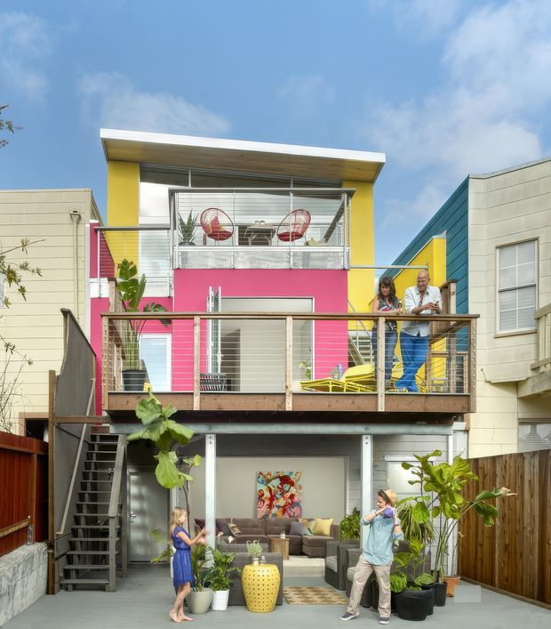 Bright, colourful multi-level urban residential facade with stacked balconies and terraces. The contemporary townhouse features vivid pink and yellow painted sections, grey and cream cladding, horizontal metal balcony railings, timber decking, exterior stairs, large windows, and an open ground-level living area. Potted tropical plants, outdoor seating, and residents add a lively, playful character.