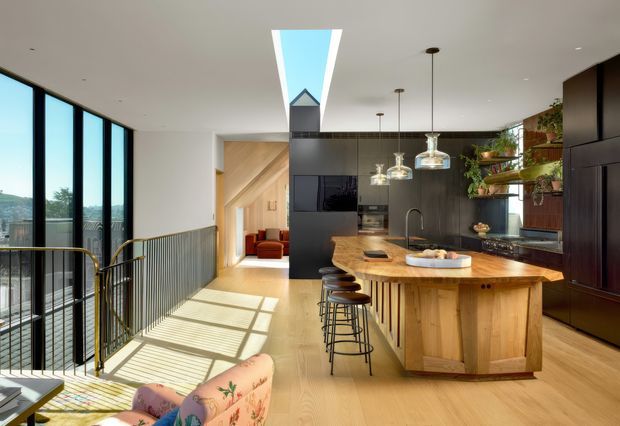 A bright, contemporary open-plan kitchen viewed from an adjoining living area or landing. The space features a large natural timber island with integrated seating, dark built-in cabinetry, a dark tiled or stone splashback, pendant lights, timber flooring, extensive floor-to-ceiling glazing, and a skylight. A black metal balustrade borders the adjacent stair or void, while plants and warm timber finishes add a natural, inviting character.