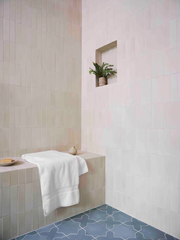 A serene, minimalist bathroom clad in pale blush and cream square tiles, with a tiled built-in bathtub or ledge extending along the corner. A white towel hangs over the edge, while a small recessed wall niche holds a potted plant. The floor features a blue and white geometric tile pattern. The space is softly lit and has a calm, refined spa-like atmosphere.