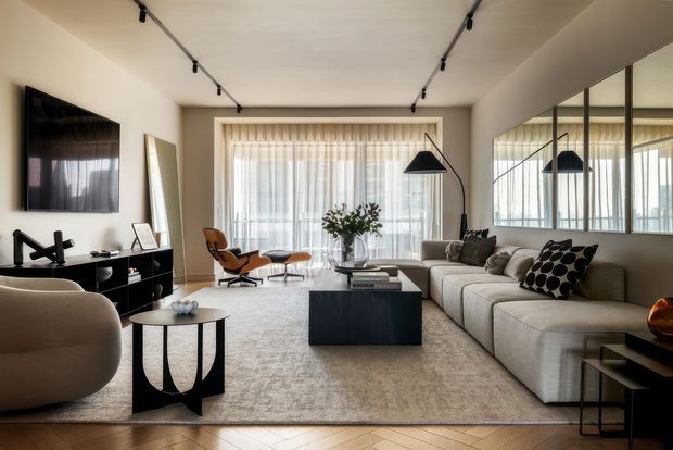 A sophisticated contemporary living room with a large cream sectional sofa, black rectangular coffee table, textured neutral rug, sculptural accent chairs, and a curved floor lamp. Full-height sheer curtains frame a bright window, while a wall-mounted television, low black media console, and large mirrored wall panels add to the polished, modern interior. Track lighting runs across the ceiling, and the timber floor is laid in a herringbone pattern.
