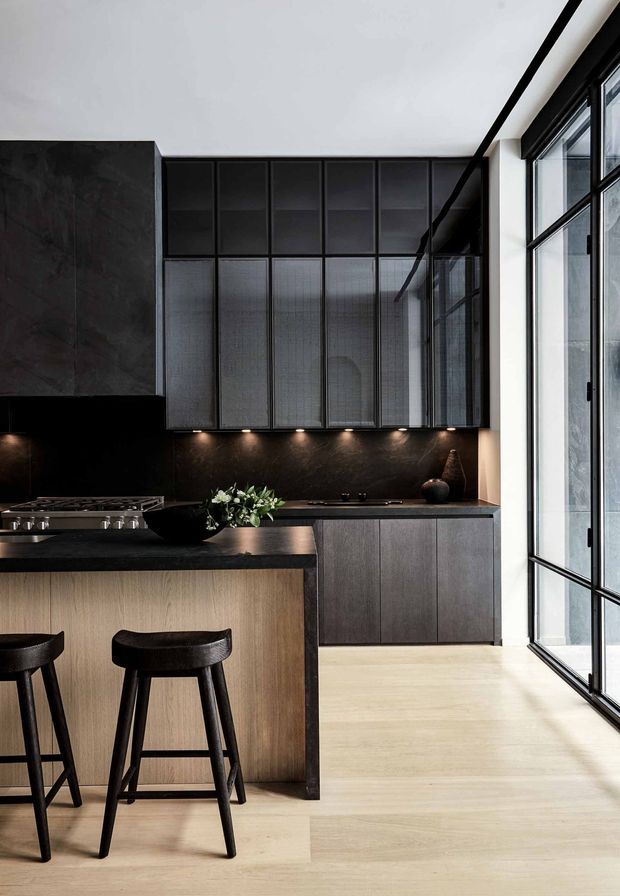 A sophisticated modern kitchen with a dark stone island and backsplash, integrated dark cabinetry, black-framed glazed panels, and a light timber island front and floor. Two black bar stools sit at the island, while full-height industrial-style glazing brings in natural light. The space has a restrained, high-contrast palette and refined contemporary detailing.