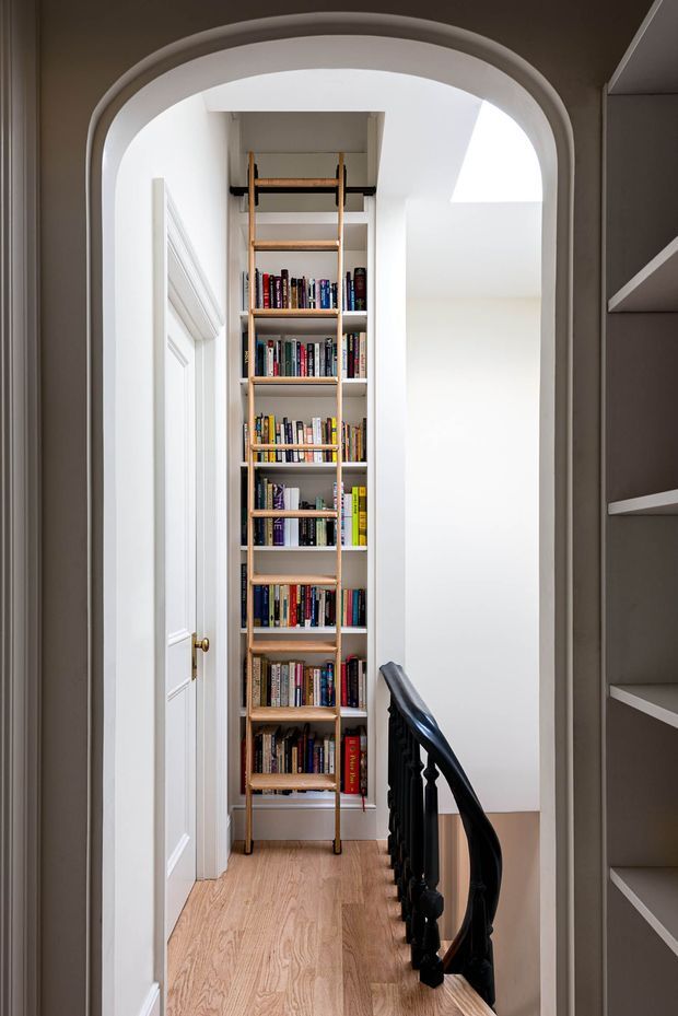 A narrow, bright residential landing or hallway framed by a large white arched opening. A tall built-in timber bookshelf fills the far wall and includes a rolling library ladder. A white paneled door sits to the left, while a dark turned timber stair balustrade and built-in shelving appear on the right. The space has light timber flooring, white walls, and strong daylight from a skylight or upper opening.