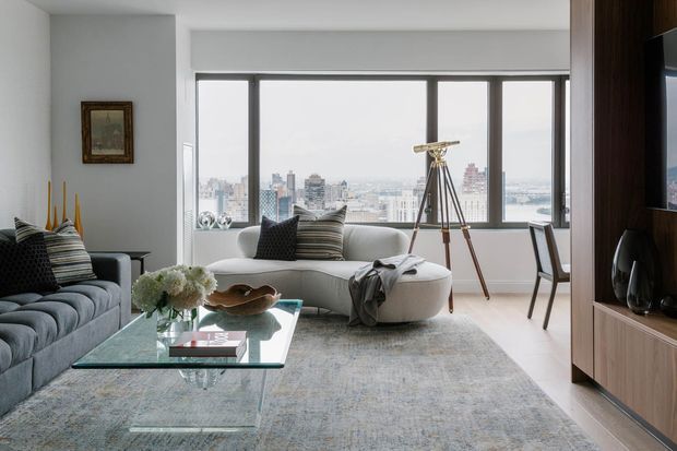 Bright, refined high-rise living room with panoramic city views through large black-framed windows. The room features a tufted grey sofa, curved cream chaise or ottoman seating, a glass coffee table, a large pale rug, sculptural decor, a telescope, and dark timber-toned built-in media cabinetry. The palette is predominantly soft grey, white, cream, and muted blue-grey, creating a calm contemporary atmosphere.