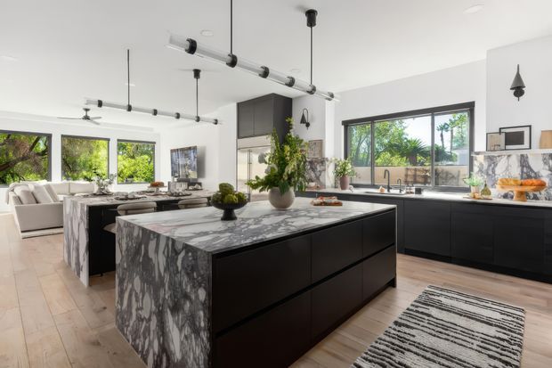 A bright, open-plan contemporary kitchen with a large dark island, dramatic veined stone waterfall benchtop, black cabinetry, and extensive garden-facing glazing. The space includes a casual dining or living area, polished concrete-style exposed services and pendant lighting, pale timber flooring, and a monochrome striped rug.