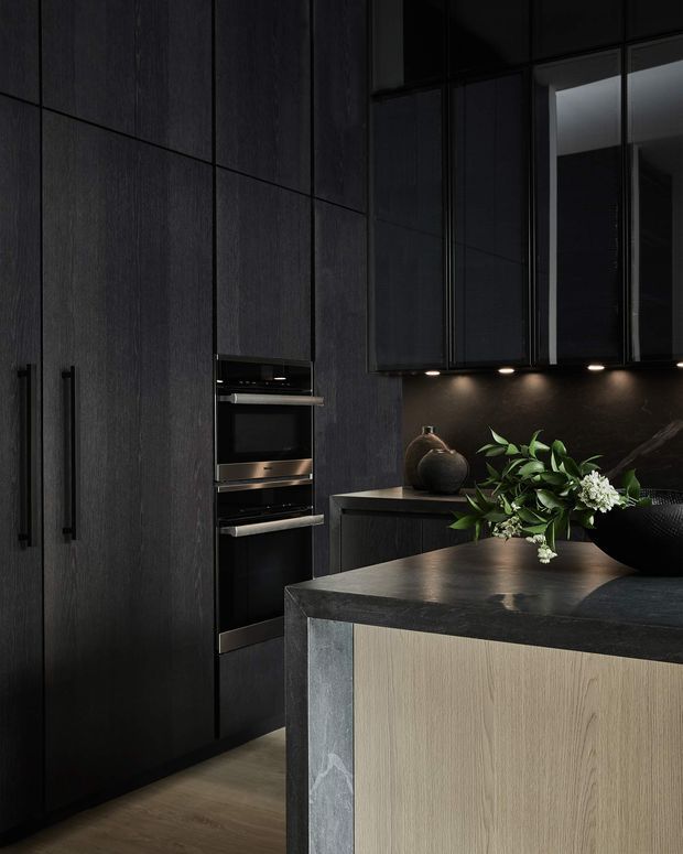 A dramatic, moody contemporary kitchen with full-height dark timber-look cabinetry, integrated black appliances, under-cabinet lighting, and a large island. The island combines a dark stone benchtop with a light timber-toned front panel and dark stone side, creating strong tonal contrast. A vase and leafy floral arrangement sit on the island.