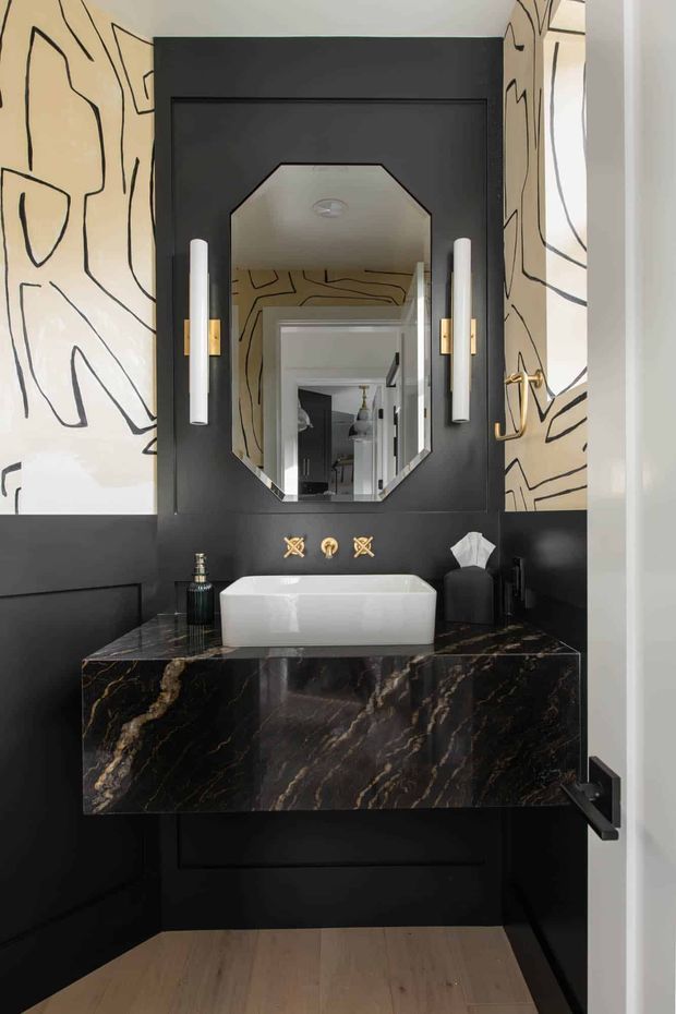 A compact, dramatically styled powder room featuring a floating black vanity with a dark veined stone benchtop, white vessel basin, brass wall-mounted tapware, and a faceted octagonal mirror. Black wall panelling is paired with cream wallpaper featuring bold abstract black linework, while slim vertical wall lights flank the mirror. Light timber flooring and a black door complete the high-contrast scheme.