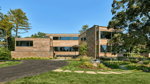A large contemporary multi-wing residence set within a landscaped suburban or wooded site. The home features extensive horizontal timber cladding, sections of exposed masonry or stone veneer, broad dark-framed glazing, and predominantly flat or very low-pitched rooflines. A curved asphalt driveway and stepping-stone garden path lead through established lawns and planting toward the building.