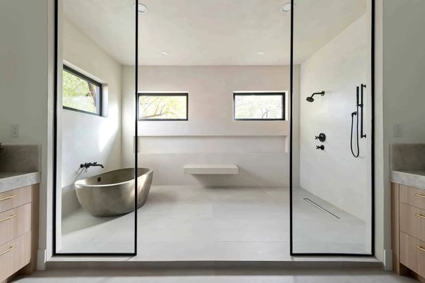 Bright contemporary bathroom viewed through a large frameless glass shower enclosure. The space features a freestanding oval bathtub, wall-mounted shower fittings, a floating bench, pale tiled or plastered surfaces, narrow horizontal windows, and timber-toned vanity cabinetry visible at both sides.
