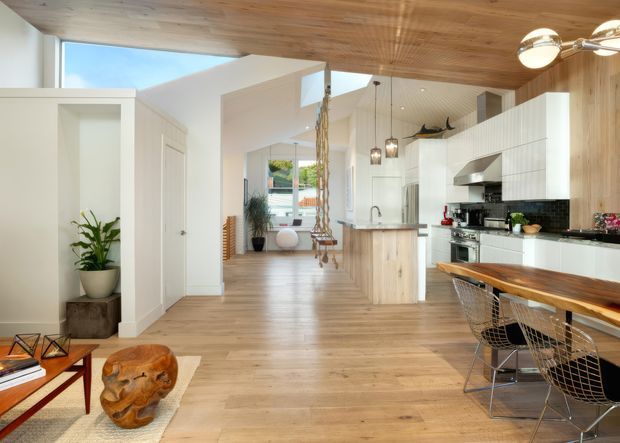 Open-plan contemporary kitchen and dining area with a warm timber-lined ceiling and matching timber flooring. White full-height cabinetry, a central island, dark tiled splashback, stainless-steel appliances, and a timber dining table with wire-frame chairs define the space. The room opens toward a bright living area and includes a visible hanging swing.