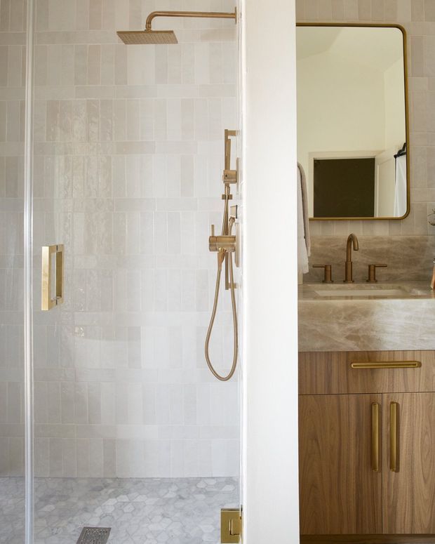 A refined bathroom featuring a glass-enclosed walk-in shower with pale rectangular wall tiles, a light marble mosaic shower floor, and brushed brass shower fittings. Beside it is a timber vanity with brass handles, a pale stone benchtop, brass tapware, and a rounded-rectangle mirror with a brass frame. The palette is warm, light, and understated.