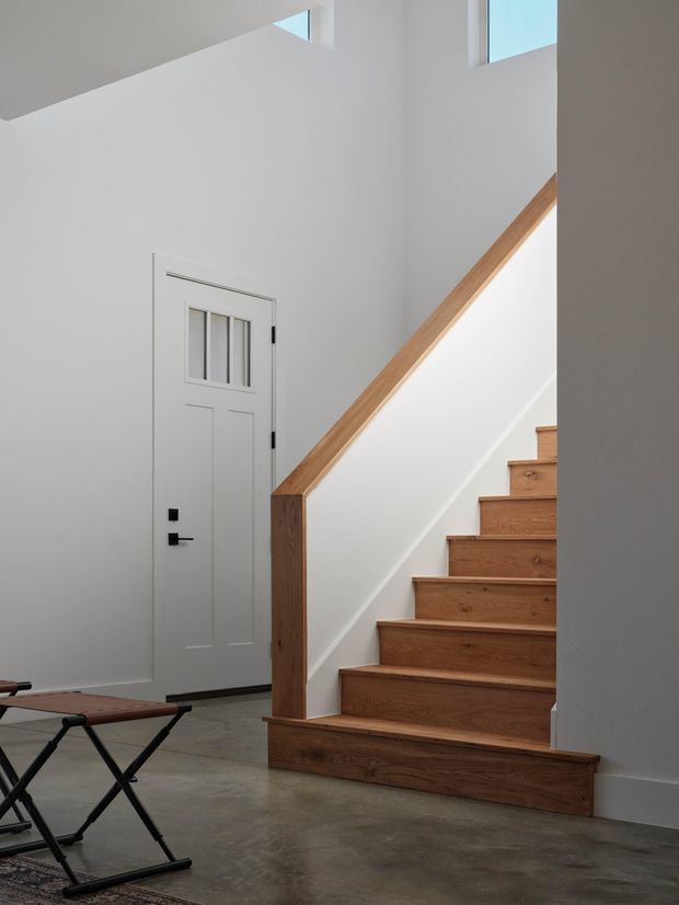 Bright minimalist interior view of a stairway and entry hall. A warm timber staircase with matching solid handrail rises beside a white wall, with a simple white panel door featuring a narrow glazed upper section. The space has polished concrete flooring, white walls, tall clerestory-style windows, and sparse black-and-leather furniture in the foreground.