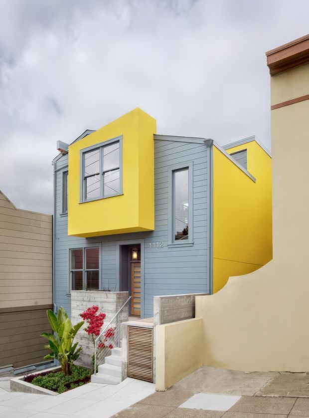 Exterior street-level view of a narrow contemporary multi-storey townhouse, featuring pale blue-grey horizontal weatherboard cladding, a vivid yellow projecting upper-storey bay and matching side wall, grey-framed windows, concrete entry walls and stairs, and a small planted frontage. The facade is tightly framed between neighbouring buildings under an overcast sky.