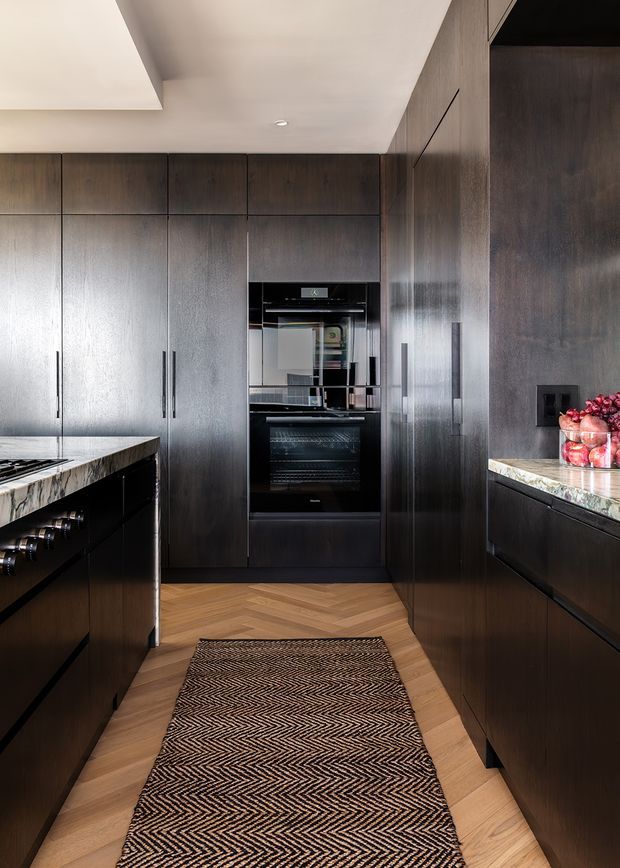 A refined contemporary kitchen with full-height dark timber cabinetry, integrated stainless-steel appliances, and a central oven stack. Pale veined stone benchtops frame the kitchen, while a warm timber herringbone floor and patterned runner soften the otherwise dark, minimalist palette. The space includes a large island and concealed storage, with recessed ceiling lighting and a clean, handle-based joinery design.