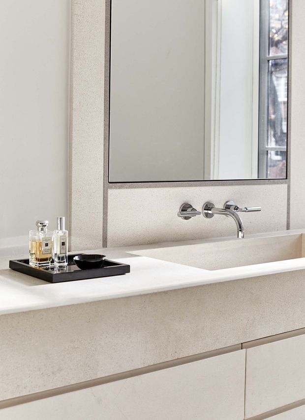 Close-up view of a refined minimalist bathroom vanity featuring a pale stone or engineered-stone benchtop and integrated basin, wall-mounted chrome tapware, a large frameless mirror, and light-coloured built-in cabinetry. A small black tray with perfume bottles sits on the counter.