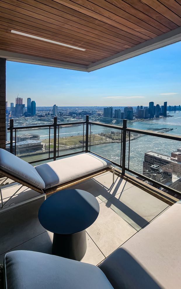 A high-rise apartment balcony or terrace overlooking a dense waterfront city skyline and river. The space has a covered timber-lined ceiling with an integrated linear light, dark structural trim, clear glass balustrades with dark metal framing, large pale tiled flooring, and contemporary outdoor lounge furniture including a cushioned chaise, chairs, and a round dark side table. The scene is bright and airy with expansive urban and water views.