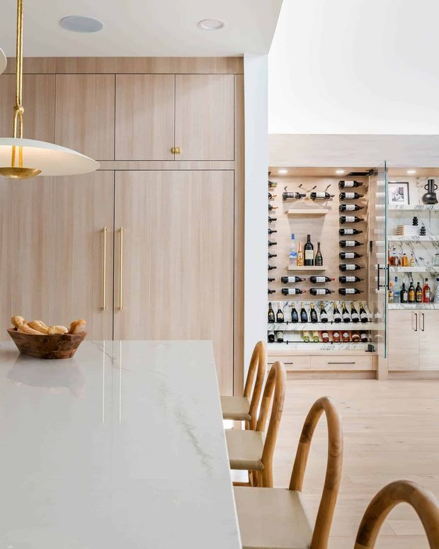 Bright contemporary kitchen and dining area with pale timber cabinetry, a large white stone island, curved timber dining chairs, brass pendant lighting, and a prominently displayed built-in wine storage wall with glass shelving and beverage cabinets.