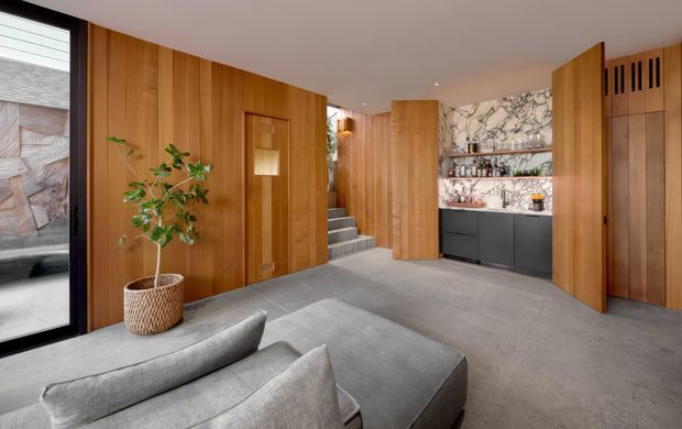 A refined contemporary entry and living area featuring extensive warm timber wall panelling and matching flush doors, a grey upholstered sectional sofa in the foreground, polished grey flooring, a short stairway, and a compact built-in bar or drinks cabinet with dark lower cabinetry and a dramatic marble-look stone backsplash. Black-framed glazing and a potted tree add contrast, while concealed lighting and clean-lined joinery create a calm, luxurious atmosphere.