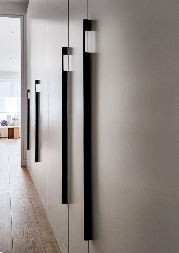 A minimalist hallway with full-height white built-in cabinetry featuring long black vertical pulls and white inset handles. Pale timber flooring runs toward a bright adjacent room, with a simple light grey wall and clean-lined contemporary detailing.