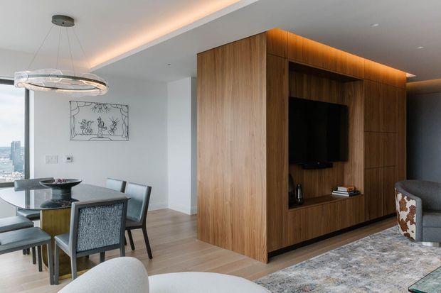 A refined open-plan living and dining area in a high-rise apartment, featuring a large walnut-toned timber entertainment wall with built-in shelving and television, pale timber flooring, a grey patterned rug, contemporary dining furniture, and a sculptural pendant light. Floor-to-ceiling glazing at left provides expansive city views, while concealed warm LED lighting adds a soft glow to the joinery and ceiling perimeter.