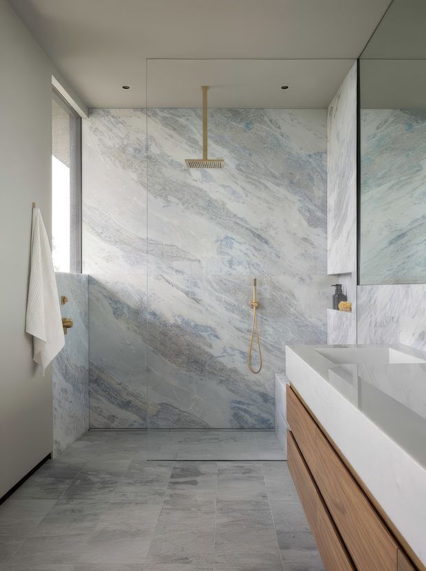 A refined contemporary bathroom featuring a frameless glass walk-in shower, full-height white and pale blue-grey veined stone wall surfaces, matching large-format floor tiles, a wall-mounted rainfall shower in a warm metallic finish, and a long white vanity with light timber cabinetry. A mirrored wall cabinet and narrow window provide additional light and reflect the stone finish.
