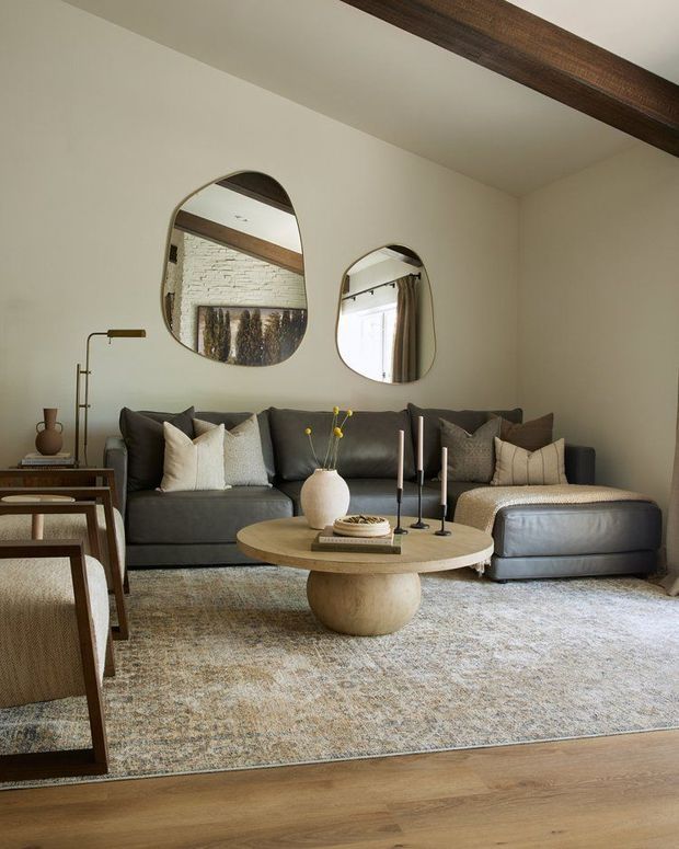 Warm, refined living room with a large dark grey sectional sofa, neutral textured cushions, sculptural round timber coffee table, tall candles, patterned area rug, and a pair of organic-shaped wall mirrors. Cream walls, exposed dark timber ceiling beam, and glimpses of brickwork and curtains in the mirror reflections add character and texture.