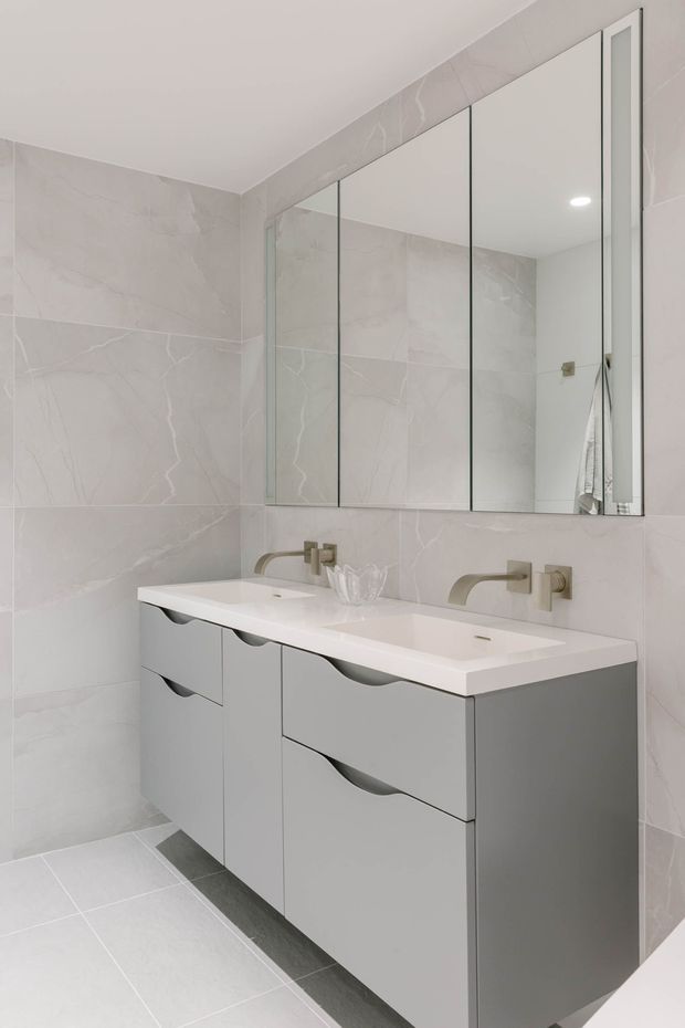 A bright, contemporary bathroom featuring a wall-mounted double vanity with integrated white basins, curved brushed-metal tapware, and grey handleless drawers. Large pale grey marble-look wall tiles continue across the room, complemented by light floor tiles. A full-width mirrored cabinet above the vanity visually enlarges the space, while recessed ceiling lighting provides even illumination.