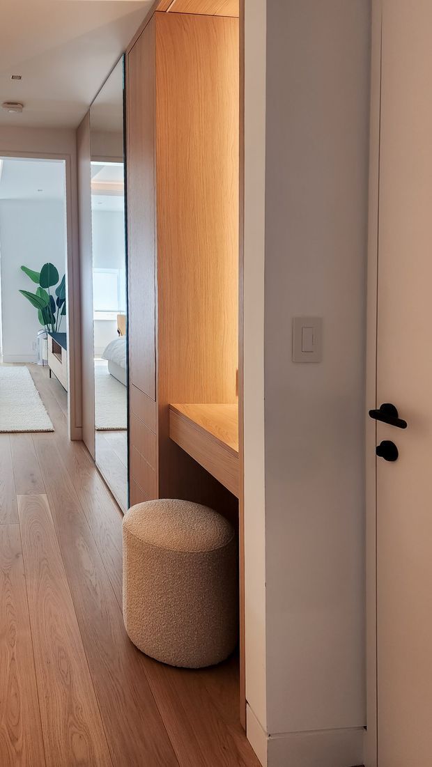 A narrow, refined residential hallway featuring full-height mirrored wardrobe or storage panels, warm timber built-in cabinetry with an integrated desk niche, and a rounded upholstered stool. Light timber flooring, soft off-white walls, and a white door with black hardware create a clean, contemporary interior. A bedroom is partially visible through the opening and reflected in the mirrors.