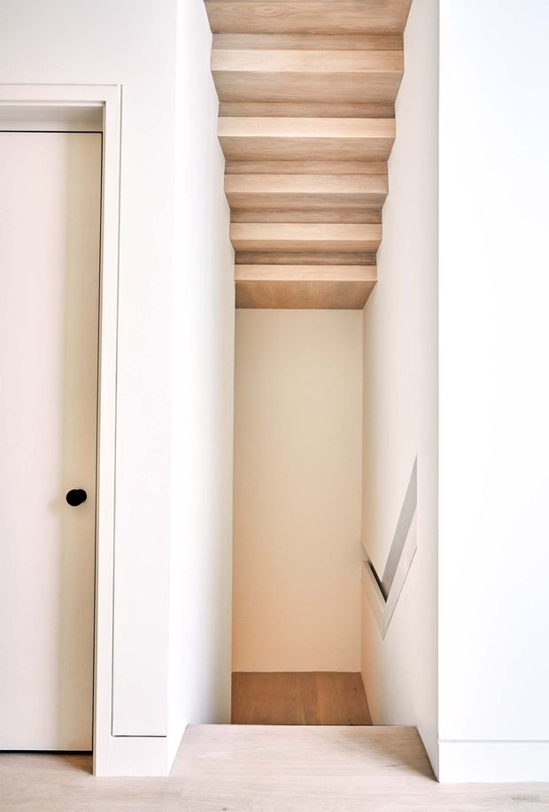Minimalist interior view of a narrow stairway descending between white plaster walls. The stair treads, lower landing, and ceiling soffit are clad in light natural timber, with a simple white flush door and dark round knob visible to the left. A slim angular handrail is integrated along the right wall. The space is bright, clean, and highly pared back.