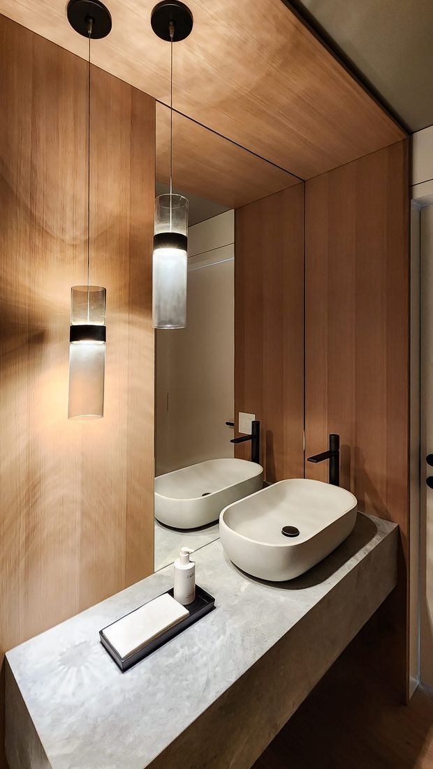 A compact, refined bathroom vanity featuring twin white vessel basins on a pale stone benchtop, matte black wall-mounted taps, warm timber wall and ceiling panelling, a large mirror, and two suspended cylindrical pendant lights. The space has a warm, contemporary hotel-like aesthetic with integrated detailing and dark timber flooring.