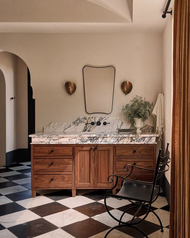 A refined bathroom vanity area with a richly grained dark timber cabinet, dramatic white marble benchtop and integrated backsplash, aged metal wall sconces, sculptural mirror, and bold black-and-white checkerboard flooring. An arched opening, black trim, and a black leather-and-metal chair add strong architectural and decorative character. Warm neutral walls and timber curtains create an elegant, vintage-inspired atmosphere.