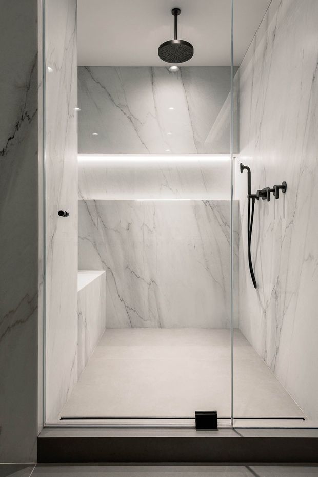 A minimalist walk-in shower enclosed by clear glass panels, finished almost entirely in large-format white marble-look slabs with grey veining. The shower includes a built-in bench, recessed illuminated niche, ceiling-mounted round rain shower, and black handheld shower fittings. The image is tightly framed on the shower enclosure and has a clean, luxurious, monochromatic appearance.