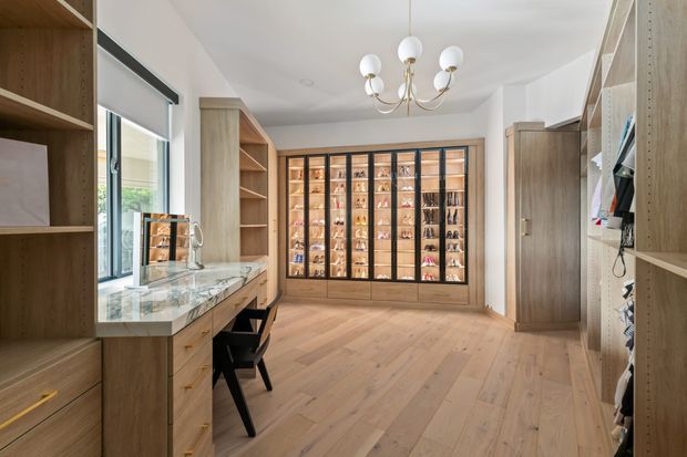 A bright, spacious walk-in wardrobe and dressing room with extensive built-in timber cabinetry, open shelving, drawers, and a large glazed display cabinet for shoes and accessories. A marble-look dressing counter sits beside the window, paired with a black chair. The room has pale timber flooring, white walls, a decorative multi-arm chandelier, and a warm, refined contemporary feel.