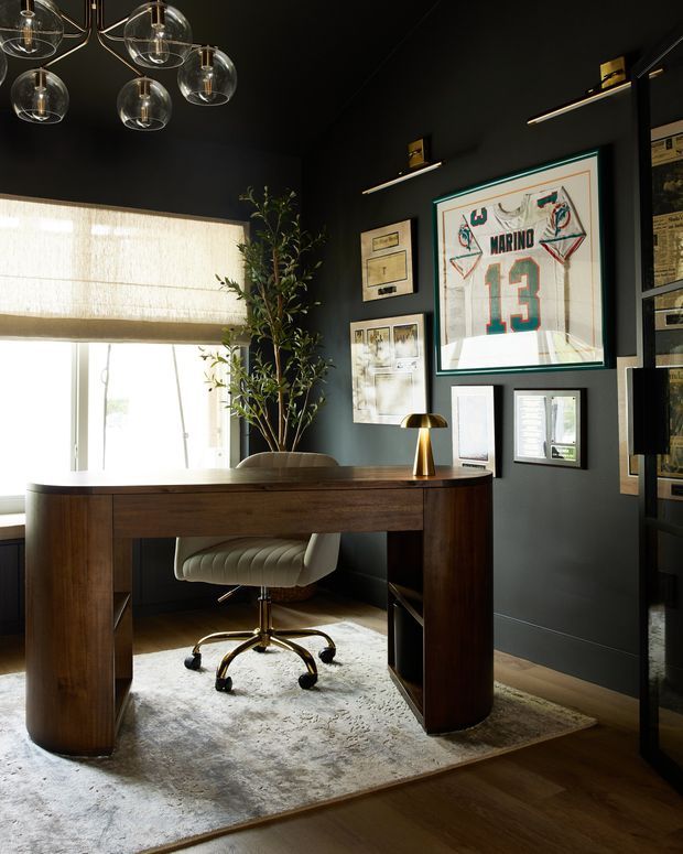 A sophisticated home office with a dark painted interior, warm timber flooring, and a substantial dark wood desk positioned in front of a large window with a woven Roman blind. The room includes a cream upholstered swivel chair, textured pale rug, brass desk lamp, indoor tree, framed sports memorabilia and artwork, and a sculptural multi-globe pendant light. The overall design is moody, refined, and masculine with warm metallic accents.