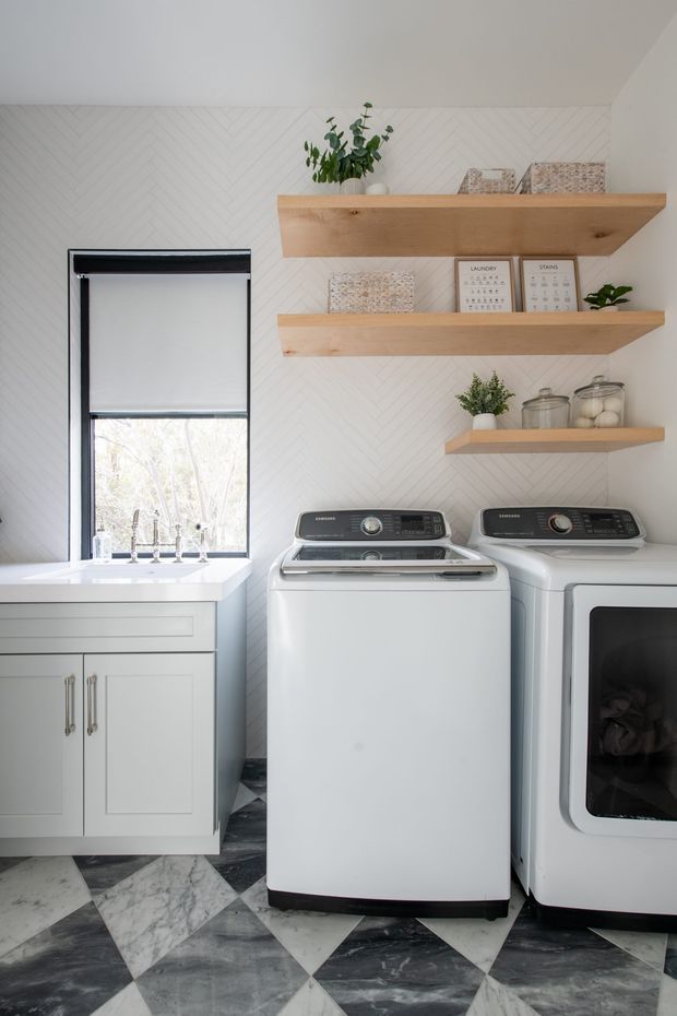 Bright, compact laundry room with a white utility sink and paneled cabinetry beside a front-loading washer and dryer. The room features white herringbone wall tiling, open light timber floating shelves with plants, framed laundry-themed prints and storage jars, plus a bold geometric marble-look floor.