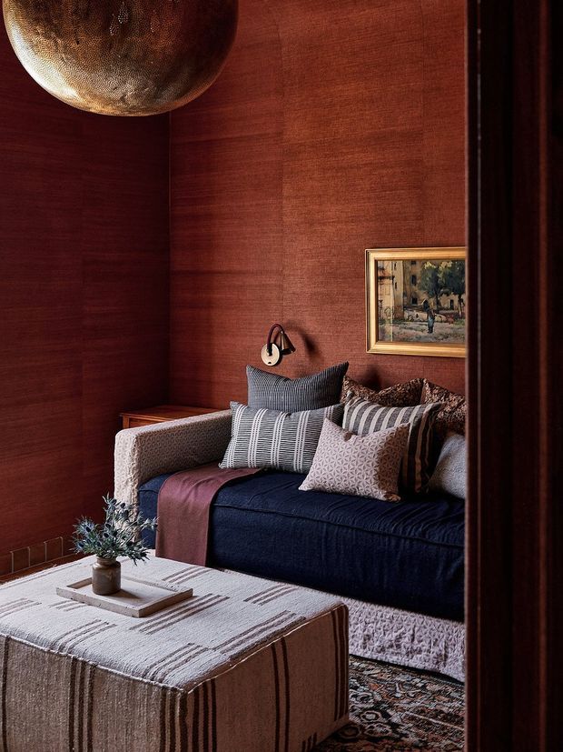 A richly layered, intimate living room centered on a deep navy sofa with patterned cushions and a rust-coloured throw. Dark reddish-brown wood or timber wall panelling creates a warm enveloping backdrop, complemented by a textured upholstered ottoman, patterned rug, framed artwork, sculptural pendant light, and a small coffee-table arrangement. The room has a moody, refined, eclectic character.
