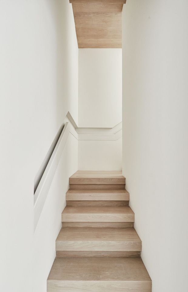 Minimalist interior photograph of a narrow staircase rising through a white-painted stairwell. The stairs and upper ceiling/landing are finished in pale natural timber, with crisp white walls, integrated simple handrails, and clean geometric detailing. The composition emphasizes the vertical void, light, and restrained material palette.