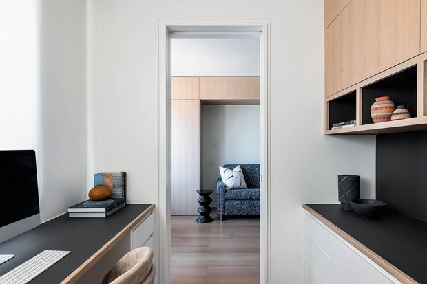 A compact, refined home office with a dark built-in desk and cabinetry, pale grey walls, and light timber detailing. A computer monitor and keyboard sit on the left workstation, while the right side features white drawers, a dark benchtop, black backsplash, and light timber overhead cupboards with open display shelving. Through the doorway, a patterned sofa and timber flooring are visible in an adjacent living area.