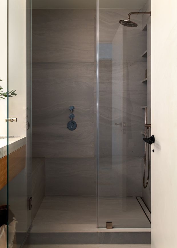 A contemporary walk-in shower enclosed by clear glass panels and door. The shower walls and floor are clad in large-format grey-beige stone-look tiles with subtle horizontal veining. Dark metal fixtures include a round overhead shower, handheld shower, controls, and linear drain. A timber-toned vanity edge is visible at left, while recessed shower shelving appears on the right.