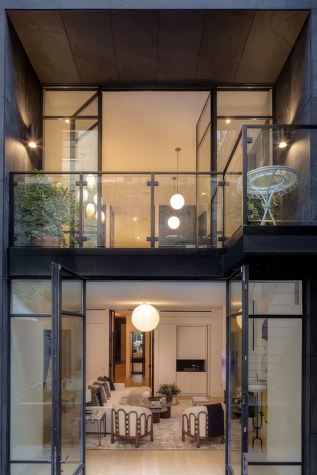 A vertically framed view into a refined multi-level residential interior through large black-framed glazed doors. The composition features an upper-level glass balustrade, dark stone or slate-like wall surfaces, a warm bronze-toned soffit, geometric glazing, and spherical pendant lights. The lower level opens into a softly furnished living room with pale walls, a large patterned rug, curved timber-framed armchairs, a round coffee table, and a sculptural fireplace or wall recess. The space has a dramatic yet warm, contemporary character with strong indoor-outdoor visual connections.