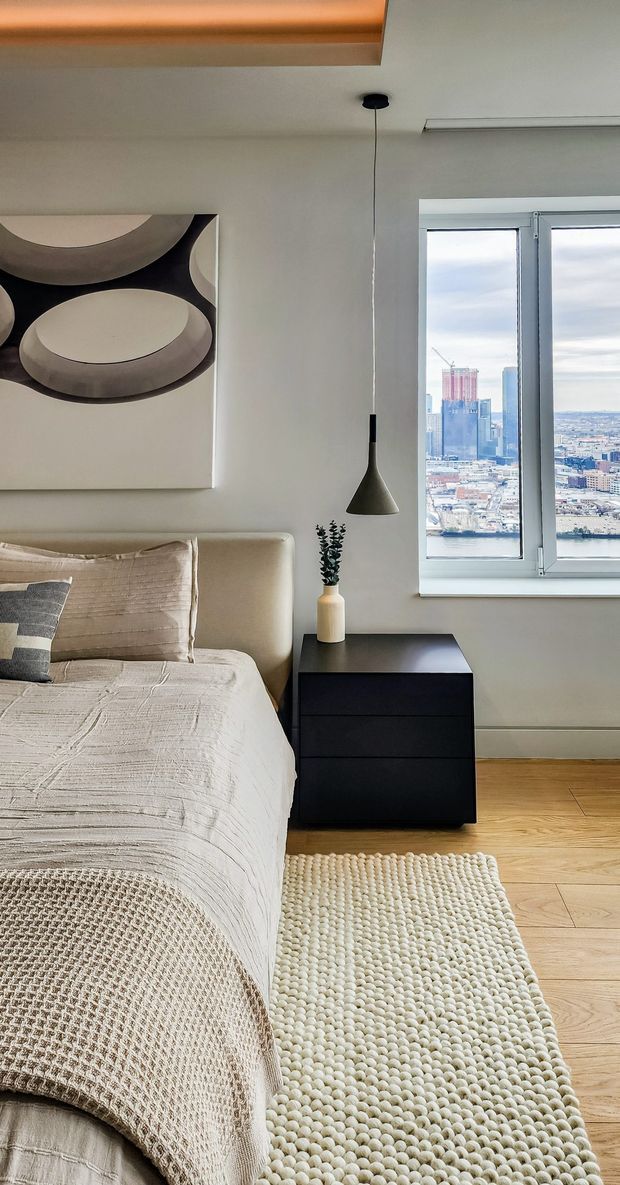 A contemporary bedroom with a neutral palette, upholstered bed, textured cream bedding, black bedside cabinet, sculptural abstract wall art, and a slim pendant light. A large window provides a city and waterfront view, while light timber flooring and a textured looped rug add warmth and softness.