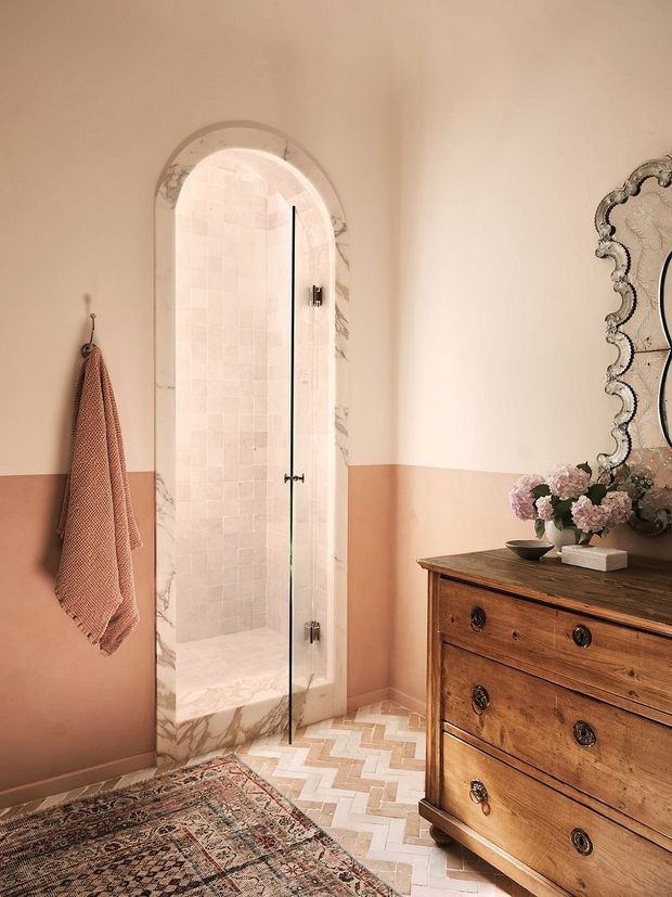 A warm, elegantly styled bathroom featuring an arched walk-in shower with a frameless glass door, pale marble-look tile, and a herringbone floor. The room has two-tone peach and cream walls, a vintage wooden chest of drawers, an ornate scalloped-edge mirror, a patterned runner rug, and floral decor.