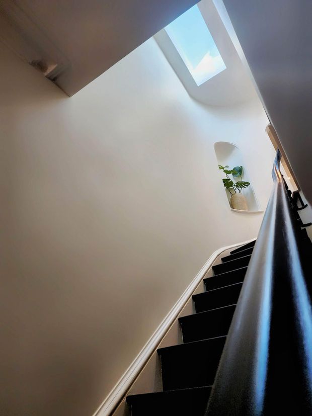 A dramatic upward view along a narrow interior staircase with dark stair treads and a glossy dark handrail, set against smooth white walls. A skylight floods the stairwell with natural light, while a small recessed shelf holds a potted plant. The composition emphasizes the vertical stairwell and strong contrast between black detailing and white surfaces.