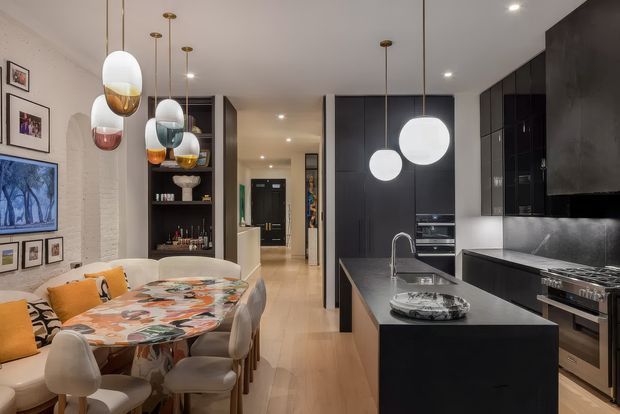 Open-plan contemporary kitchen and dining area with a dark, full-height kitchen wall and central island, adjoining a round dining table surrounded by upholstered seating. Glossy black cabinetry, dark stone surfaces, integrated appliances, light timber flooring, white textured walls, and sculptural pendant lights create a refined, high-contrast interior.