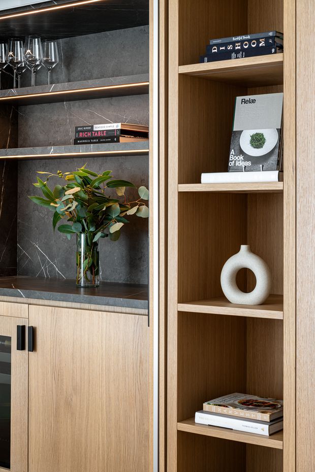 Close-up interior detail of bespoke built-in shelving and cabinetry. The joinery combines warm light timber veneer with dark grey marble- or stone-look backing and shelving, integrated linear LED lighting, black hardware, books, wine glasses, and understated decorative objects. The composition has a refined, contemporary built-in display and bar-storage character.