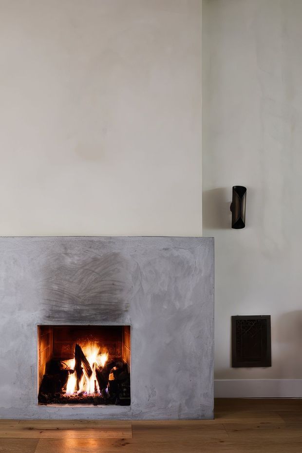 Interior detail of a minimalist fireplace with a live wood fire, set into a broad smooth grey plaster or concrete surround. The room has pale warm-toned walls, timber flooring, a small black wall light, and a dark ventilation grille, creating a restrained, warm and atmospheric composition.