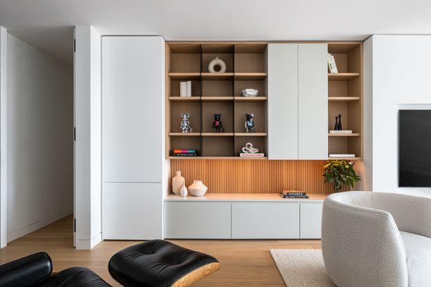 A refined contemporary living room centred on a full-width built-in timber and white cabinetry wall. Open shelving displays decorative objects and books above a fluted timber feature backsplash and low white storage units, with a television partially visible at right. The room has warm timber flooring, a neutral upholstered lounge chair, a black leather lounge chair and ottoman, and soft natural light.