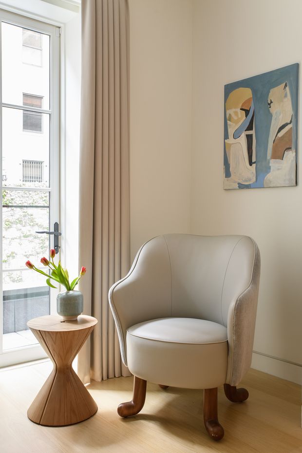 A bright, refined interior corner featuring a sculptural upholstered armchair beside a small hourglass-shaped timber side table with a ceramic vase and flowers. Full-height beige curtains frame a glazed door or window, while a muted abstract artwork hangs on the warm off-white wall. Pale timber flooring and soft neutral furnishings create a calm, airy atmosphere.